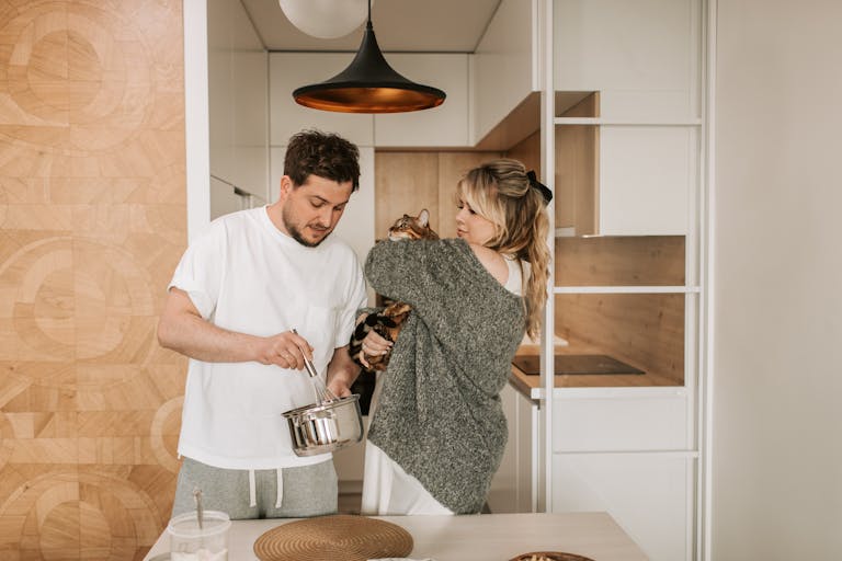 A couple with a cat enjoy cooking together in a stylish, minimalist kitchen, showcasing love and warmth.